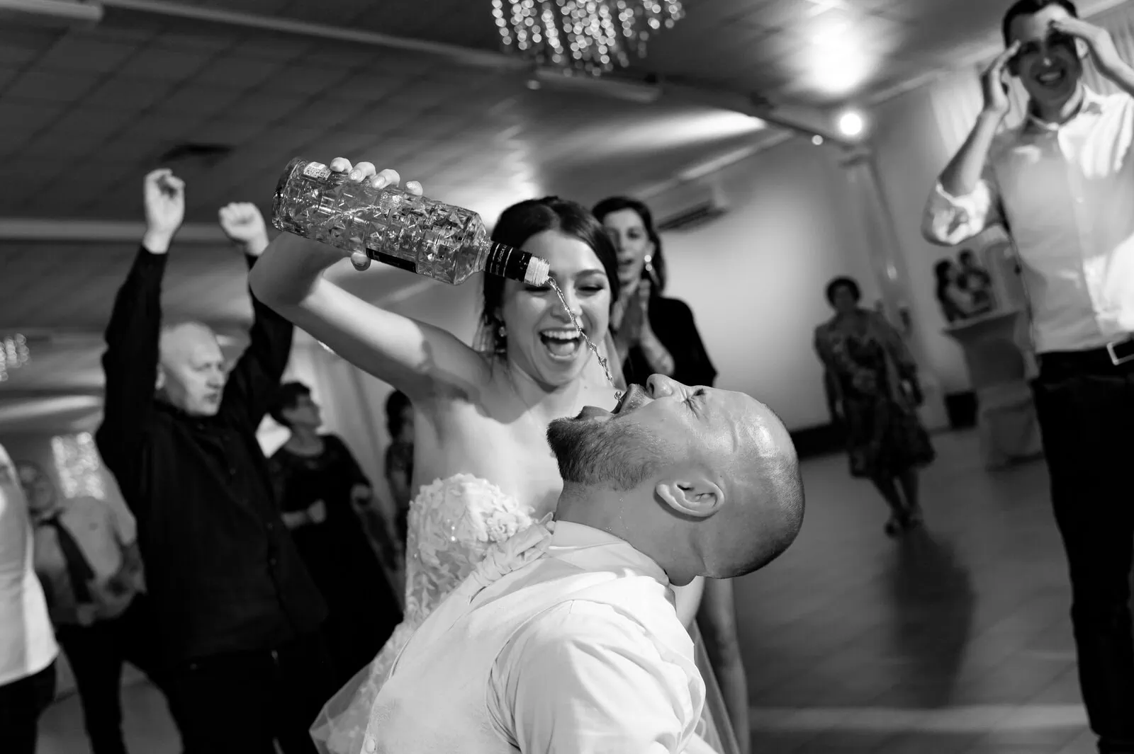 Florale Hochzeitsfeier mit tanzender Braut, die Wasser in den Mund eines lachenden Bräutigams gießt, umgeben von tanzenden Gästen, schwarz-weiße Aufnahme, festliche Stimmung, Hochzeitsevent, Hochzeitsfotografie.