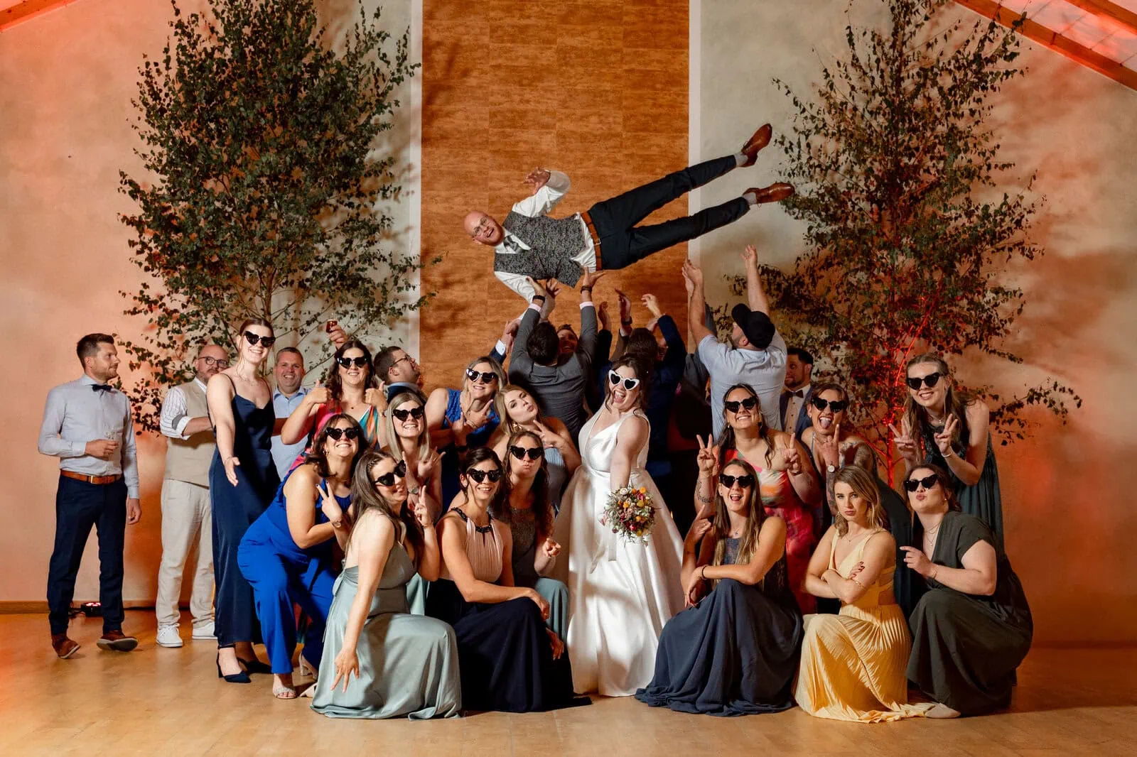 Junges Brautpaar mit Freunden bei der Hochzeitsfeier, alle tragen Sonnenbrillen, lachen und posieren für eine lebendige Hochzeitsgruppenaufnahme.